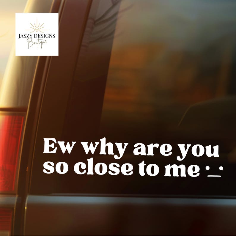 May include: A white vinyl decal applied to a dark car window featuring the text "Ew why are you so close to me :_" in a bold, rounded font. A small logo in the top left corner displays the text "JASZY DESIGNS Boutique" with a sun icon.