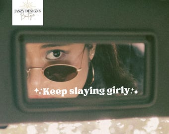 May include: A rectangular car visor mirror featuring the white text Keep slaying girly with four-pointed star icons on either side. The mirror reflects a portion of a face wearing oval-shaped sunglasses and large hoop earrings.