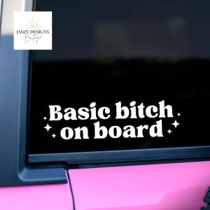 May include: White vinyl decal with the text "Basic bitch on board" in a bold, retro font, accented with starbursts. The decal is displayed on a black car window, with a pink car panel visible.