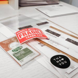 May include: A collection of shipping materials including a red and white sticker with the text HANDLE WITH CARE FRAGILE THANK YOU, a Curad alcohol prep pad, a circular black sticker with the text THANK YOU, and various printed order documents.