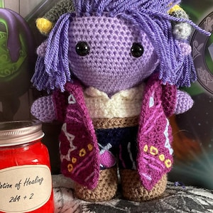 Op de afbeelding: Handgemaakte gehaakte pop met paars haar en een bijpassende outfit. De pop heeft een wit shirt, een donkerblauw vest en een magenta jasje met gele en lichtblauwe accenten. Een potje rode vloeistof met het label "Potion of Healing" staat voor de pop.