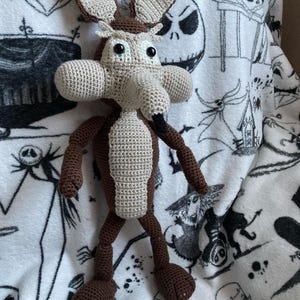 May include: A handmade crochet figure of Wile E. Coyote, featuring a tan and brown color scheme. The cartoon character has large ears, a long snout, and black eyes. The plush toy is resting on a white blanket with a black and white print.