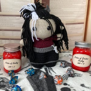 Op de afbeelding: Handgemaakte gehaakte pop met zwart en wit haar, gekleed in een bordeauxrode en grijze top en zwarte rok. Twee potten rode vloeistof met het label "Healing Potion" en dobbelstenen suggereren een tabletop-spelthema.