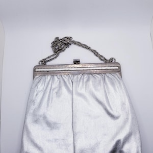 May include: Silver clutch purse with a chain strap. The purse has a rectangular frame with a clasp closure and a pleated body. The chain is a silver color. The purse is made of a shiny, textured material.