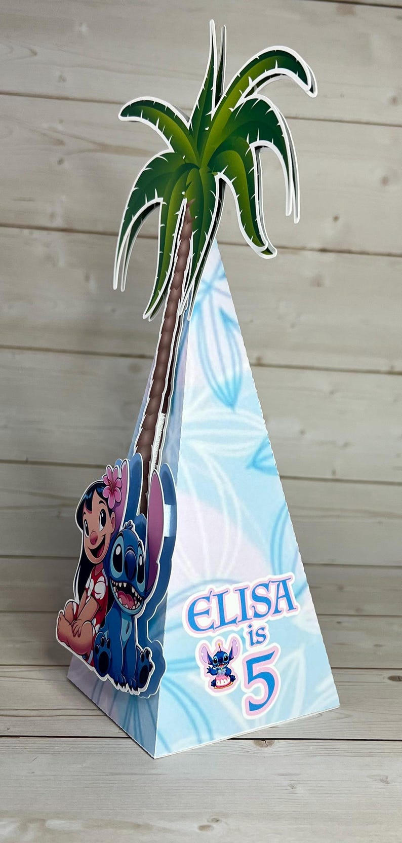 Op de afbeelding: Blauwe en witte feestversiering in de vorm van een kegel, met een palmboom en stripfiguren. De decoratie heeft de naam "Elisa" en het nummer "5" erop.