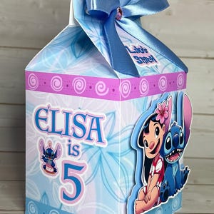 Op de afbeelding: Een lichtblauwe geschenkdoos met een bloemenpatroon en de tekst "Elisa is 5". De doos is versierd met afbeeldingen van Lilo en Stitch en een blauwe strik. Perfect voor een verjaardag.