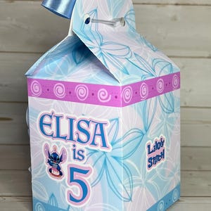 Op de afbeelding: Een gepersonaliseerde geschenkdoos met een bloemenpatroon in blauw en roze. De doos is versierd met de naam "ELISA", het nummer "5" en een stripfiguur. Een blauw lint is aan de bovenkant bevestigd.