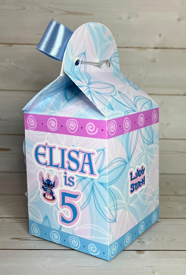 Op de afbeelding: Een gepersonaliseerde geschenkdoos met een bloemenpatroon in blauw en roze. De doos is versierd met de naam "ELISA", het nummer "5" en een stripfiguur. Een blauw lint is aan de bovenkant bevestigd.
