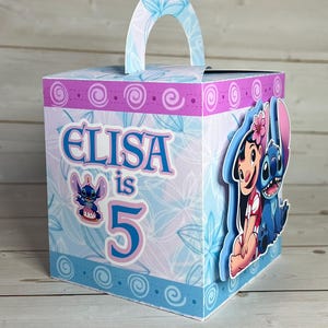 Op de afbeelding: Een gepersonaliseerde cadeauverpakking met Lilo & Stitch-figuren. De doos heeft een blauw-roze bloemenpatroon. De tekst "ELISA is 5" staat op de voorkant. Met een handvat aan de bovenkant.
