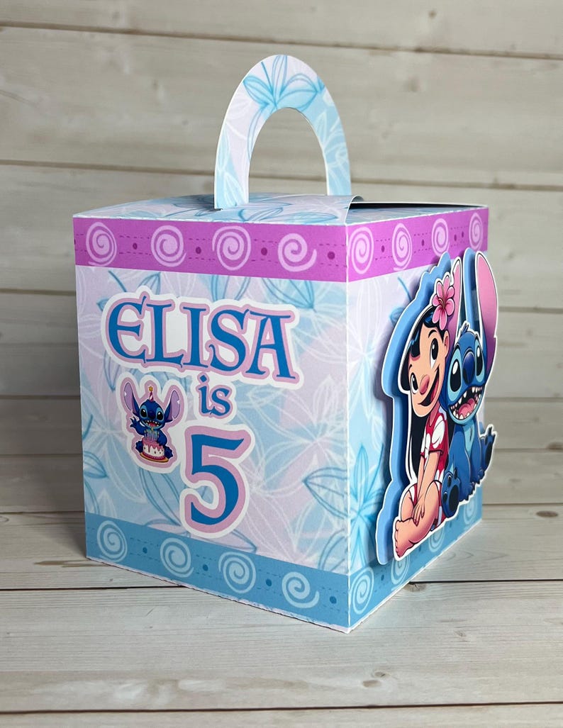 Op de afbeelding: Een gepersonaliseerde cadeauverpakking met Lilo & Stitch-figuren. De doos heeft een blauw-roze bloemenpatroon. De tekst "ELISA is 5" staat op de voorkant. Met een handvat aan de bovenkant.