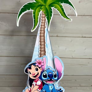 Op de afbeelding: Een decoratief kegelvormig middelpunt met een palmboom en de tekenfilmfiguren Lilo en Stitch. De palmboom heeft groene bladeren en een bruine stam. Lilo draagt een rode jurk en Stitch is blauw.
