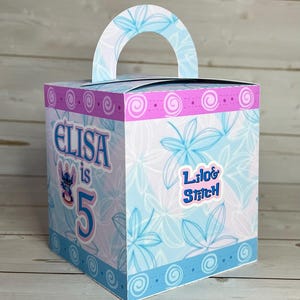 Op de afbeelding: Vierkante geschenkdoos met een handvat, versierd met een blauw-wit bloemmotief. De doos heeft de tekst "Elisa is 5" en een "Lilo & Stitch" sticker. Roze en blauwe randen sieren de boven- en onderranden.