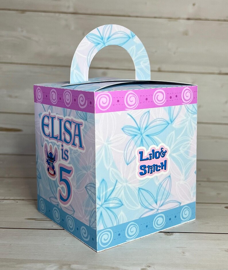 Op de afbeelding: Vierkante geschenkdoos met een handvat, versierd met een blauw-wit bloemmotief. De doos heeft de tekst "Elisa is 5" en een "Lilo & Stitch" sticker. Roze en blauwe randen sieren de boven- en onderranden.