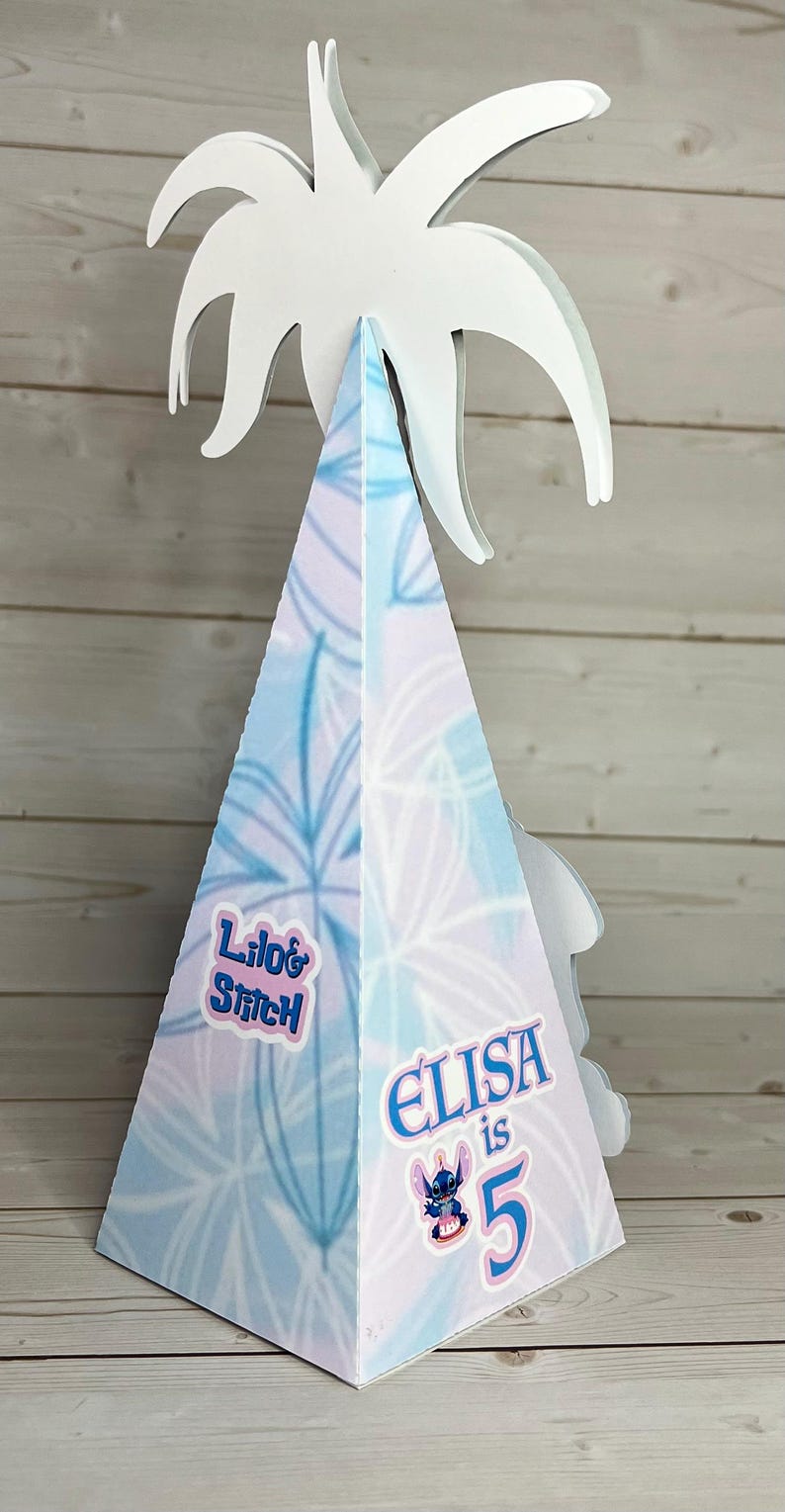 Op de afbeelding: Een kegelvormige feesthoed met een witte palmboomtop. De hoed is versierd met een blauw-wit bloemenpatroon en de tekst "Lilo & Stitch" en "Elisa is 5" met een Stitch-afbeelding.