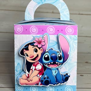 Op de afbeelding: Decoratieve geschenkdoos met de personages Lilo en Stitch. De doos heeft een lichtblauw handvat en is versierd met bloemmotieven en een roze en blauwe rand. Lilo draagt een rode jurk en Stitch is blauw.