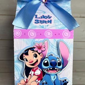 Op de afbeelding: Decoratieve geschenkdoos met Lilo & Stitch-personages. De doos heeft een blauwe strik met een roze edelsteen. De voorkant van de doos toont Lilo en Stitch, met de woorden "Lilo & Stitch" erboven. De doos heeft een lichtblauwe en witte achtergrond.