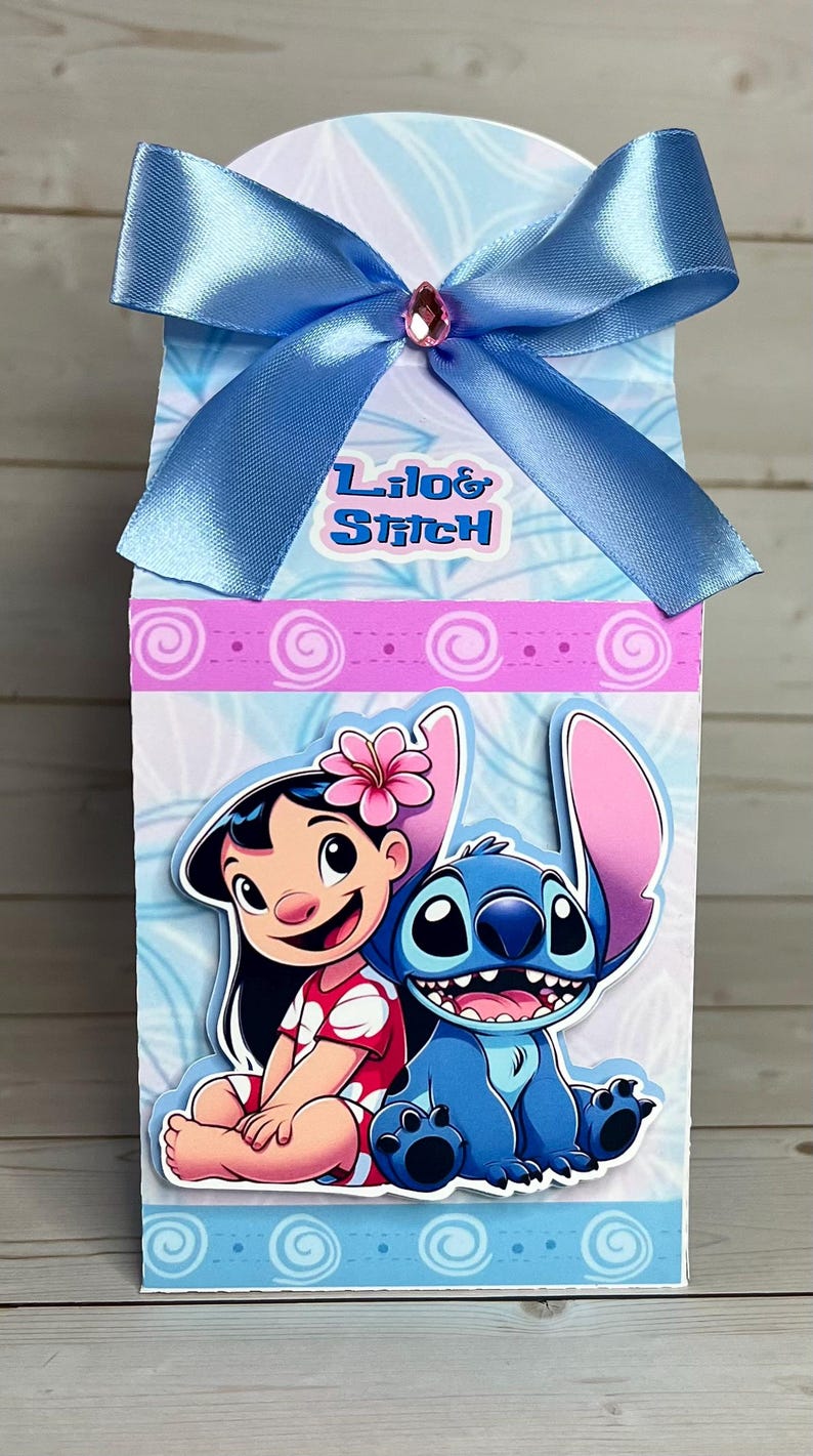 Op de afbeelding: Decoratieve geschenkdoos met Lilo & Stitch-personages. De doos heeft een blauwe strik met een roze edelsteen. De voorkant van de doos toont Lilo en Stitch, met de woorden "Lilo & Stitch" erboven. De doos heeft een lichtblauwe en witte achtergrond.