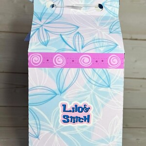 Op de afbeelding: Een hoge, rechthoekige geschenkdoos met een blauwe strik aan de bovenkant. De doos heeft een bloemenpatroon in blauw- en wittinten, met een roze band en de tekst "Lilo & Stitch" in blauw.