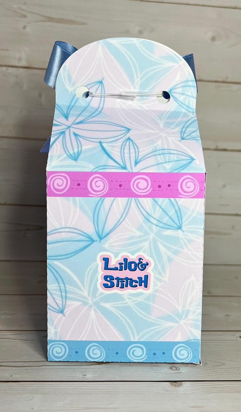 Op de afbeelding: Een hoge, rechthoekige geschenkdoos met een blauwe strik aan de bovenkant. De doos heeft een bloemenpatroon in blauw- en wittinten, met een roze band en de tekst "Lilo & Stitch" in blauw.