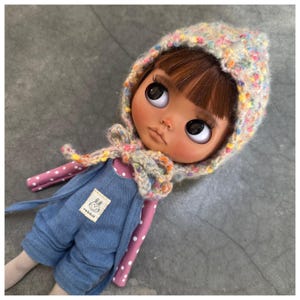 Può includere: Una bambola con capelli castani e grandi occhi che indossa un cappello di lana con macchie colorate e una tuta di jeans blu. La bambola indossa anche una camicia rosa a pois bianchi. Una piccola etichetta con un logo di coniglio è sulla tuta.