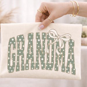 Op de afbeelding: Cr&egrave;mekleurige draagtas met het woord "GRANDMA" in groene letters met stippen. Een witte strik staat rechts van de tekst. De tas wordt vastgehouden door een hand met gouden sieraden.