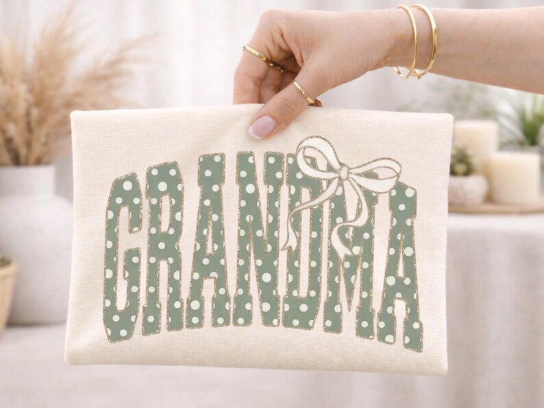Op de afbeelding: Cr&egrave;mekleurige draagtas met het woord "GRANDMA" in groene letters met stippen. Een witte strik staat rechts van de tekst. De tas wordt vastgehouden door een hand met gouden sieraden.