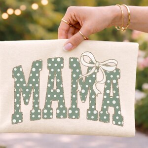 Op de afbeelding: Cr&egrave;mekleurige draagtas met het woord "MAMA" in groene stippenletters. Boven de laatste letter staat een witte strik. De tas wordt vastgehouden door een hand met gouden ringen en armbanden.
