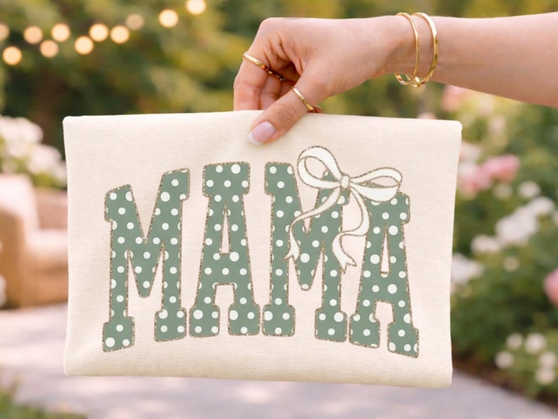 Op de afbeelding: Cr&egrave;mekleurige draagtas met het woord "MAMA" in groene stippenletters. Boven de laatste letter staat een witte strik. De tas wordt vastgehouden door een hand met gouden ringen en armbanden.
