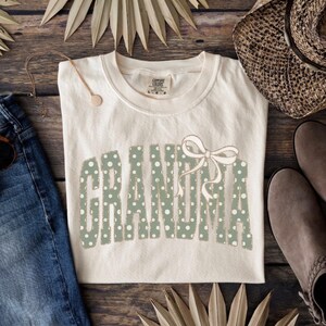 Op de afbeelding: Cr&egrave;mekleurig T-shirt met het woord "GRANDMA" in groene stippenletters en een strik. Het shirt wordt gepresenteerd op een houten oppervlak met een spijkerbroek, een ketting, een hoed en laarzen.