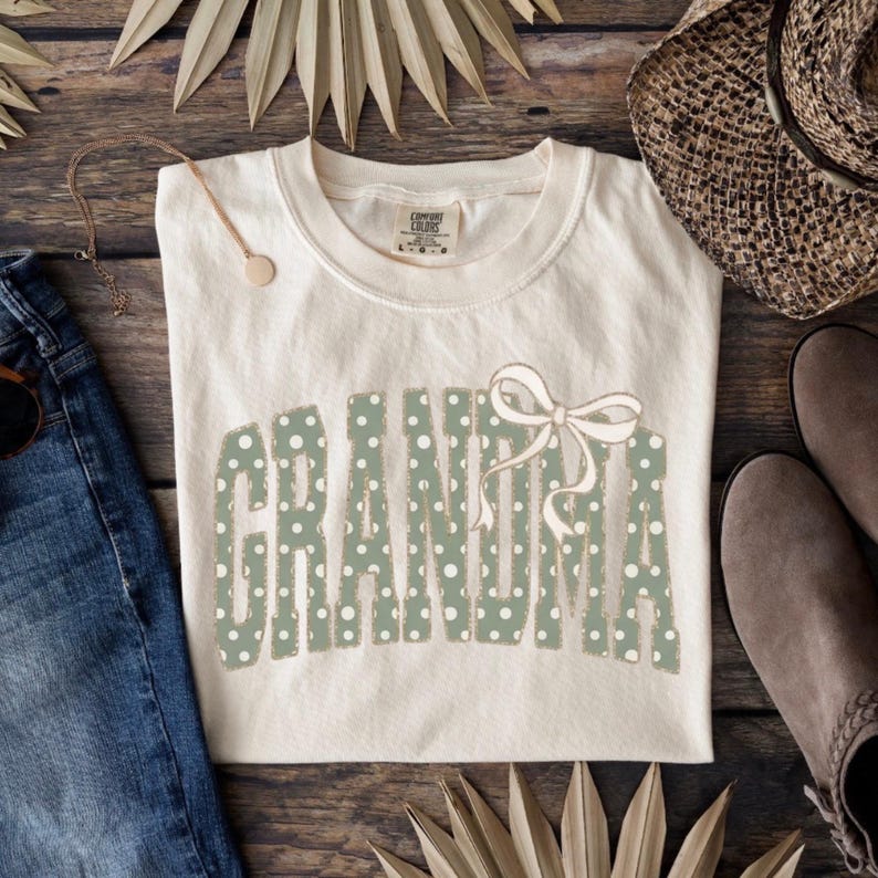 Op de afbeelding: Cr&egrave;mekleurig T-shirt met het woord "GRANDMA" in groene stippenletters en een strik. Het shirt wordt gepresenteerd op een houten oppervlak met een spijkerbroek, een ketting, een hoed en laarzen.