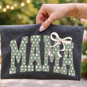 Op de afbeelding: Een donkergrijze clutch met het woord "MAMA" in grote, groen gestippelde letters. Een witte strik is boven de letter "A" geplaatst. De achtergrond is wazig.