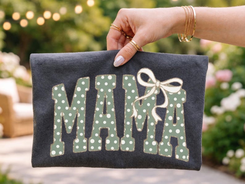 Op de afbeelding: Een donkergrijze clutch met het woord "MAMA" in grote, groen gestippelde letters. Een witte strik is boven de letter "A" geplaatst. De achtergrond is wazig.