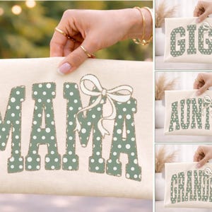 Op de afbeelding: Cr&egrave;me canvas clutch met het woord "MAMA" in groene stippenletters en een witte strik. Extra clutches met "GIGI", "AUNTIE" en "GRANDMA" worden ook getoond.