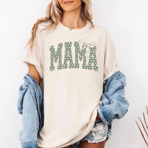 Op de afbeelding: Cr&egrave;mekleurig T-shirt met het woord "MAMA" in groene stippenletters en een bijpassende strik. Het shirt heeft een relaxte pasvorm en opgerolde mouwen. Een spijkerjasje wordt over het shirt gedragen.