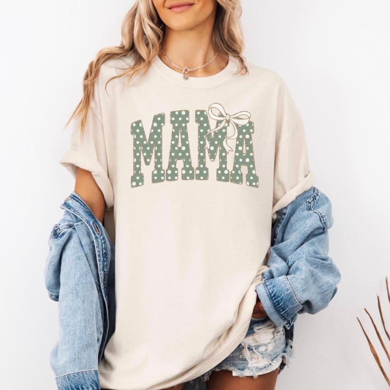 Op de afbeelding: Cr&egrave;mekleurig T-shirt met het woord "MAMA" in groene stippenletters en een bijpassende strik. Het shirt heeft een relaxte pasvorm en opgerolde mouwen. Een spijkerjasje wordt over het shirt gedragen.