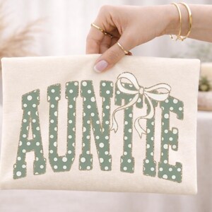 Op de afbeelding: Cr&egrave;mekleurige clutch met het woord "AUNTIE" in groene stippenletters en een witte strik. De clutch wordt vastgehouden door een persoon met gouden sieraden.