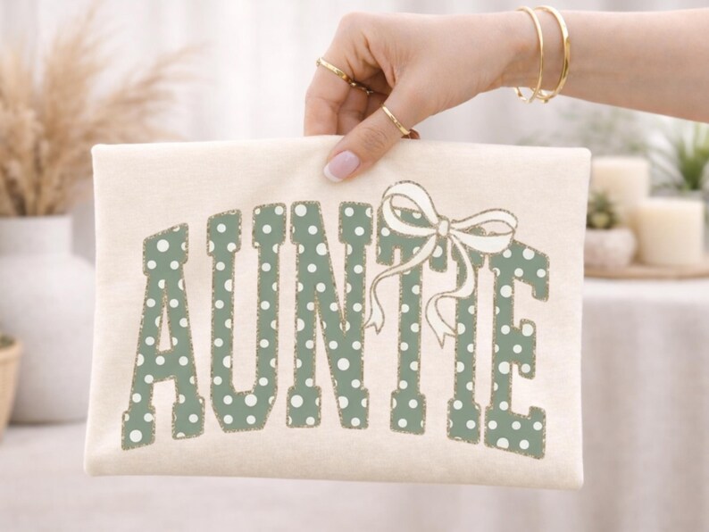 Op de afbeelding: Cr&egrave;mekleurige clutch met het woord "AUNTIE" in groene stippenletters en een witte strik. De clutch wordt vastgehouden door een persoon met gouden sieraden.