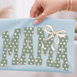 Op de afbeelding: Een lichtblauw t-shirt met het woord "MAMA" in grote, groen gestippelde letters. Een witte strik met gouden rand is boven de laatste letter geplaatst. Het shirt wordt door een hand omhoog gehouden.