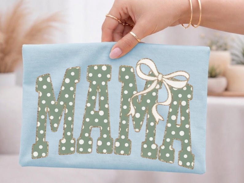 Op de afbeelding: Een lichtblauw t-shirt met het woord "MAMA" in grote, groen gestippelde letters. Een witte strik met gouden rand is boven de laatste letter geplaatst. Het shirt wordt door een hand omhoog gehouden.