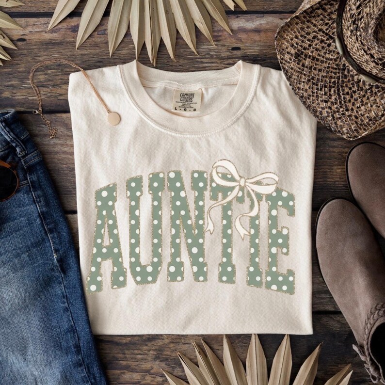 Op de afbeelding: Cr&egrave;mekleurig T-shirt met het woord "AUNTIE" in groene stippenletters en een bijpassende strik. Het shirt wordt gepresenteerd op een houten oppervlak met een spijkerbroek, een ketting, een hoed en laarzen.