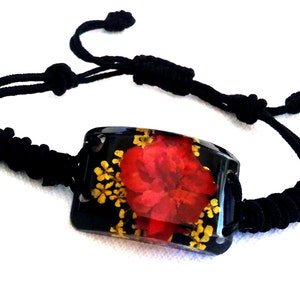 May include: Black cord bracelet with a rectangular resin charm featuring a red flower and gold accents.