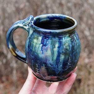 May include: A handmade ceramic mug with a unique handle design. The mug features a glossy glaze with shades of blue and green, creating a marbled effect. The mug is held in a hand, showcasing its size and shape. Perfect for coffee or tea.