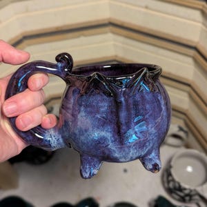 May include: A purple and blue ceramic mug shaped like a cauldron with a handle and four legs. The mug has a speckled glaze and a white interior.