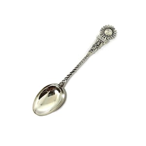 May include: A silver spoon with a twisted handle and a decorative end featuring a sun design and the text "ST.MORITZ". The spoon has a polished, reflective bowl.