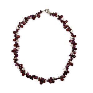 Op de afbeelding: Een ketting met een delicaat ontwerp van druppelvormige edelstenen in tinten diep rood en helder wit. De edelstenen zijn aan elkaar geregen, waardoor een vloeiende, organische vorm ontstaat. De ketting is vastgezet met een zilverkleurige sluiting.