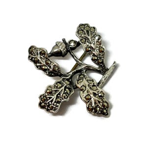 Op de afbeelding: Zilverkleurige broche in de vorm van een eiken tak met bladeren en een eikel. De bladeren zijn gedetailleerd met kleine, donkere steentjes, wat een sprankelend effect creëert. De broche staat tegen een witte achtergrond.