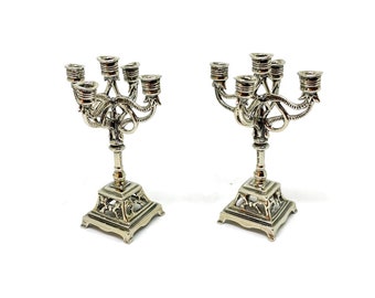 May include: Two silver menorahs with ornate designs and seven candle holders each. The menorahs have a square base with intricate details.