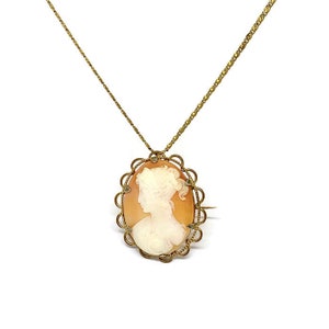 Op de afbeelding: Een goudkleurige ketting met een ovale camee hanger. De camee toont het profiel van een vrouw in wit tegen een perzikkleurige achtergrond, omlijst door een decoratieve gouden rand. De ketting is delicaat en past bij de vintage uitstraling van de hanger.