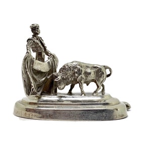 May include: A silver figurine of a matador and a bull. The matador, in a detailed outfit, holds a cape, facing the charging bull. The figurine is mounted on a round base. The sculpture captures the drama of the bullfight, with fine detailing.