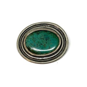 Op de afbeelding: Een ovale broche van zilver met een groene turquoise steen in het midden. De steen wordt omringd door twee concentrische cirkels van zilveren kralen.